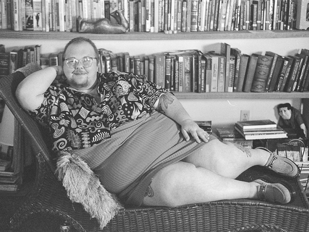 A white person with short facial hair and head hair, wearing a decorative button-down top and a tight skirt, one hand tucked behind their head and the other resting against their side.They're sitting on their side along a wicker chaise. A packed 3-tier bookshelf sits behind them.