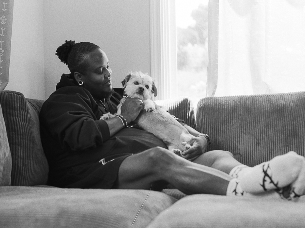 A Black person with short hair styled in an Afro puff holds a Shih Tzu against their chest, their legs up as they're nestled in the corner of a corduroy couch. They're wearing a Black hoodie, shorts, and socks with an image of barbed wire across them.