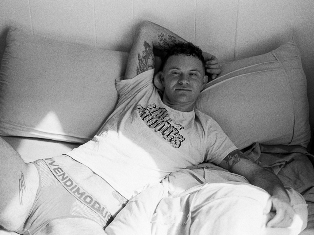 A white trans man lying diagonally across a bed, one tattooed arm bent behind his head with his fingers curled around the side of his ear. The other arm is extended out to his side, against his bedsheet. He's smiling with a closed mouth, wearing a white t-shirt with decorative lettering, and Vendimodus briefs, his legs slightly spread.