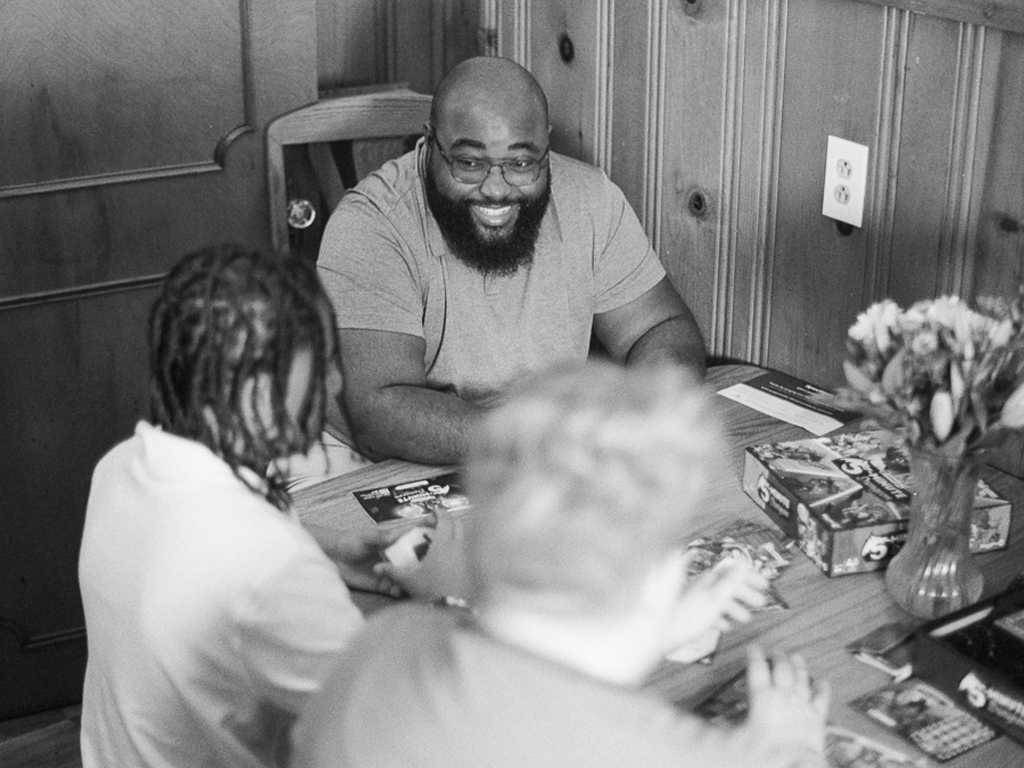 A bald Black man with a medium-length beard and glasses rests his hands on a wooden dining table, seated at the head of the table and smiling widely at 2 people seated perpendicular to him. The other peoples' backs are turned to the camera; on the left, a child wears their hair in dark locs. The person on the right has light blonde hair. The 3 people are putting together a puzzle on the table.