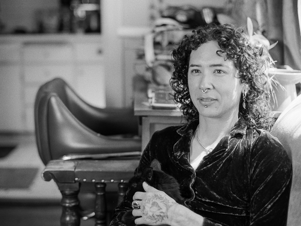 A person with shoulder-length curly brown hair and facial piercings sits in an armchair, one hand stroking a black cat in their lap. They are seated in a living room area, with a kitchen in the background. They're wearing a black, velvet collared vest.