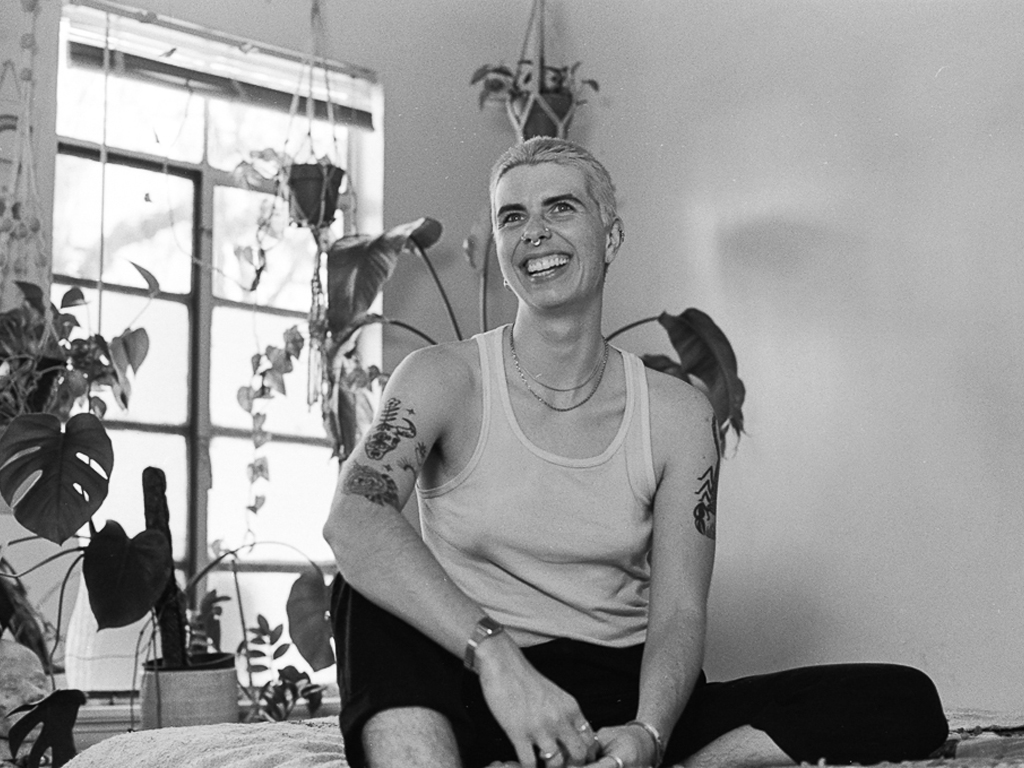 A white person with short blond hair, upper arm tattoos ,and several facial piercings, wears a white tanktop and their hands held in front of their body. Several large plants are scattered around a multi-pane window in the background, sunlight pouring through. Their smiling toothily.