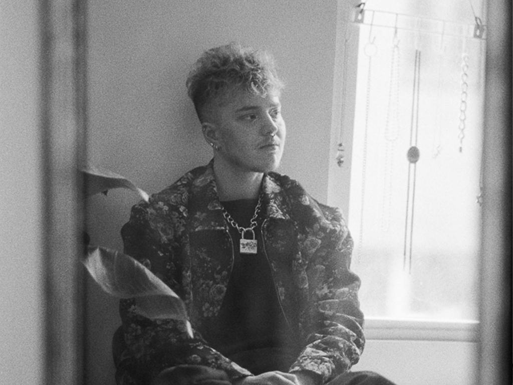 A white person with short, curly blonde hair sits on a bed, visible here in their reflection in a mirror. They're wearing a floral denim jacket and a chain necklace with a padlock on it. They're glancing off to one side, expressionless, seated next to a window with sunshine pouring in.