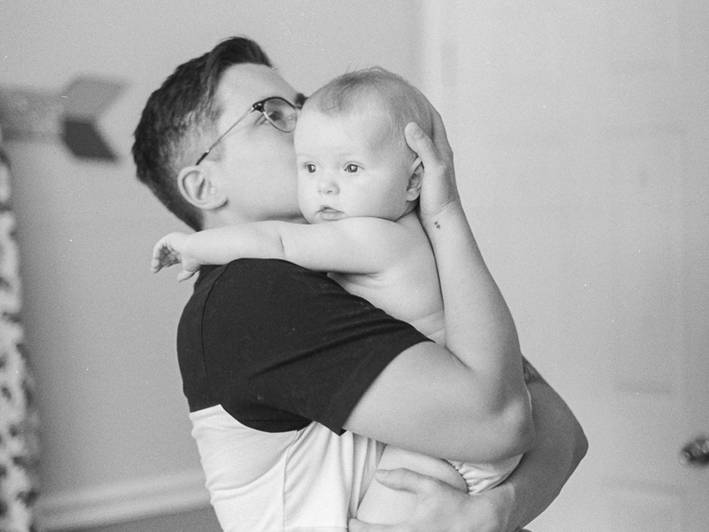 a white person holds a baby in a diaper against their chest, kissing the side of the baby's temple.