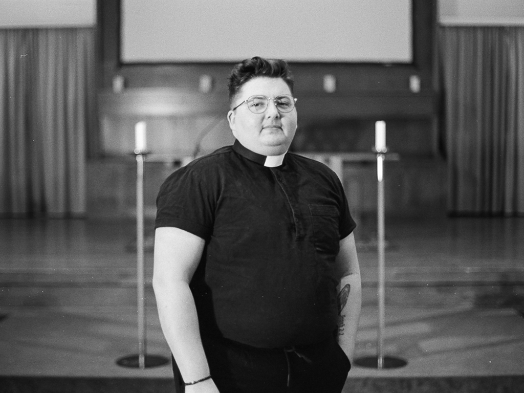 A light-skinned Mex-indigenous person stands in front of a church altar, at the end of the aisle, one hand in their pocket. They're wearing a black short-sleeve shirt with a clerical collar, short brown spiked hair, and sandals. Tall candlesticks and candles are staggered behind them