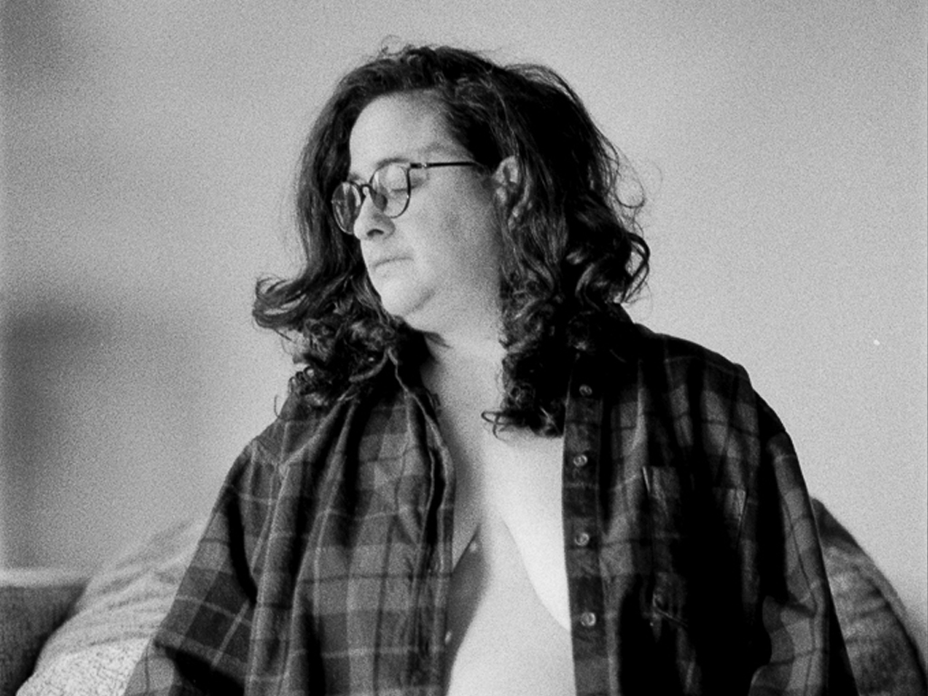 A white trans masculine person with chest-length dark brown hair sits on the edge of an armchair cushion wearing an open plaid button-down. They're wearing black Calvin Klein boxer briefs, their hands perched gently on either leg. Half-moon rimmed glasses rest on their nose as they look down to one side.
