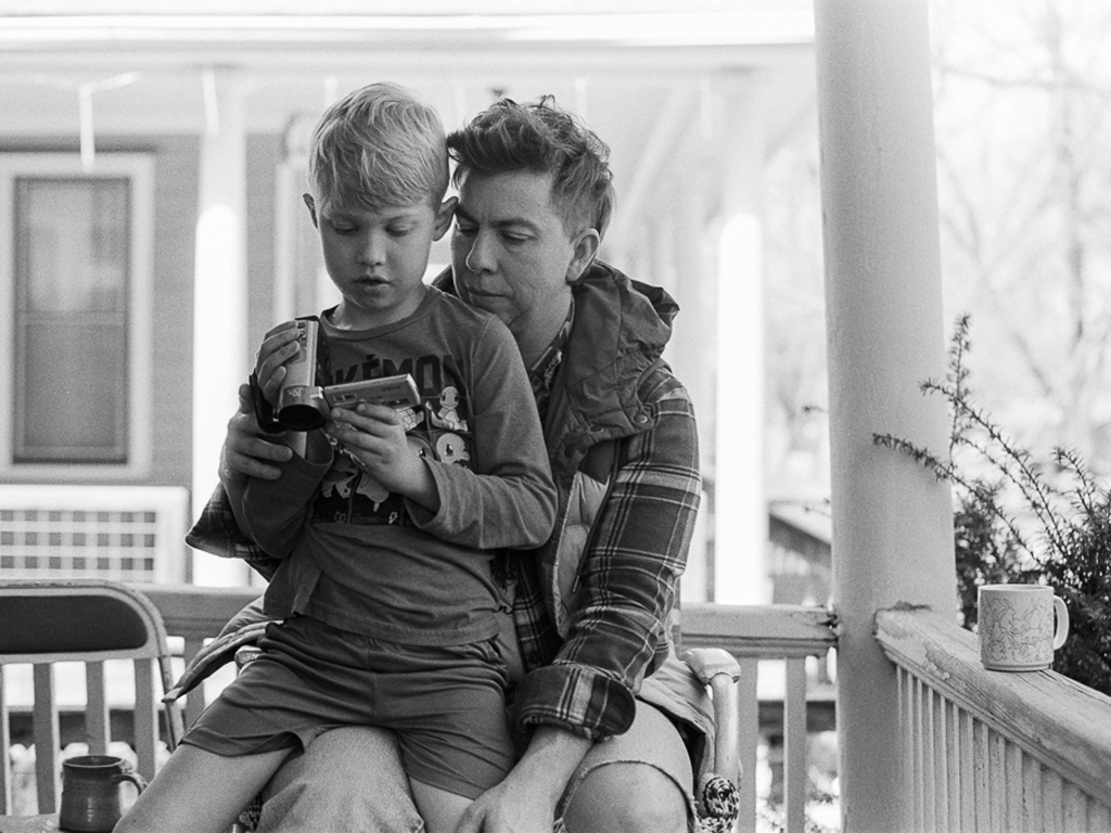 A white trans masculine person with dirty blonde hair and a jacket with plaid sleeves sits with a child in their lap. The pair are looking interestedly into the side-display of a digital video camera. They sit outdoors on the porch of a house, a railing behind them.