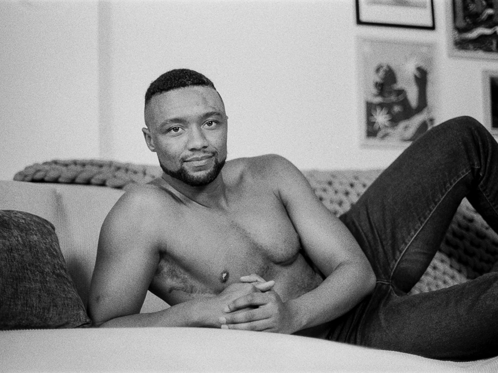 A Black man with short brown hair, shaved on the sides of his head in a fade. He has short facial hair on his chin, and is shirtless with black jeans. He's on his side across the bench of a white couch, a weighted crocheted blanket across the back. Four pieces of framed artwork line the wall behind him.